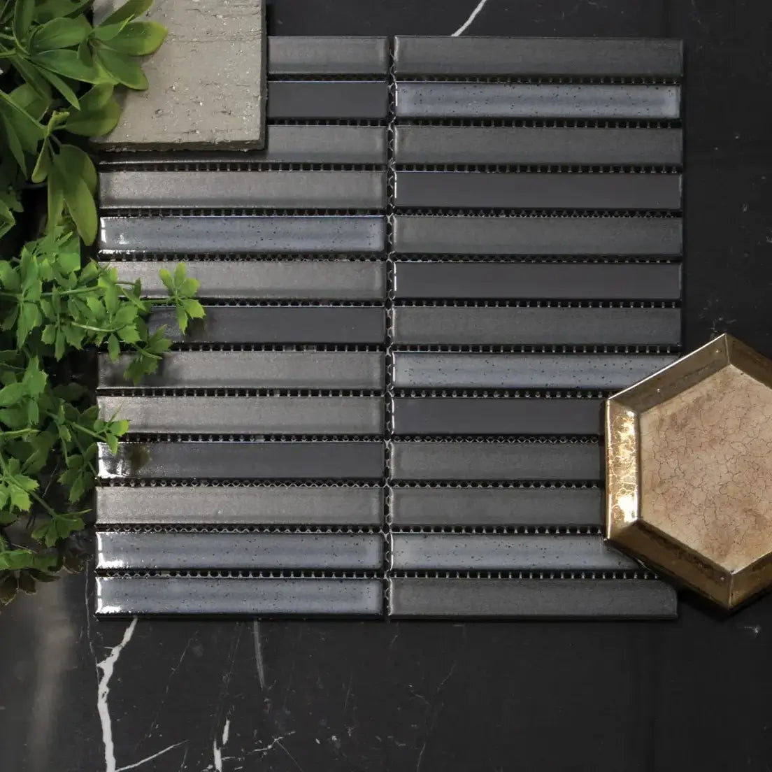 The Sevilla Mosaic Tiles, KitKat design in a Original Carbon Shade Gray Mix, with a size of 20x145mm mounted on a dark marble effect wall. Green plants leaning in from the left side.