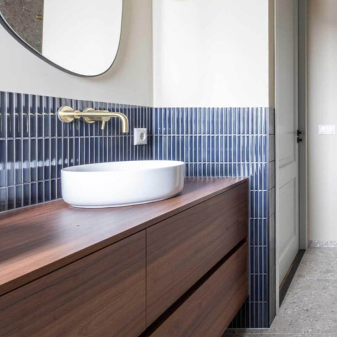 The Sevilla Mosaic Tiles, Original KitKat design in a Blue Gray Glossy, with a size of 20x145mm. Installed in a bathroom setting, a brown vanity unit with a white sink mounted on top with a gold painted tap set above and a wall mounted mirror above that.