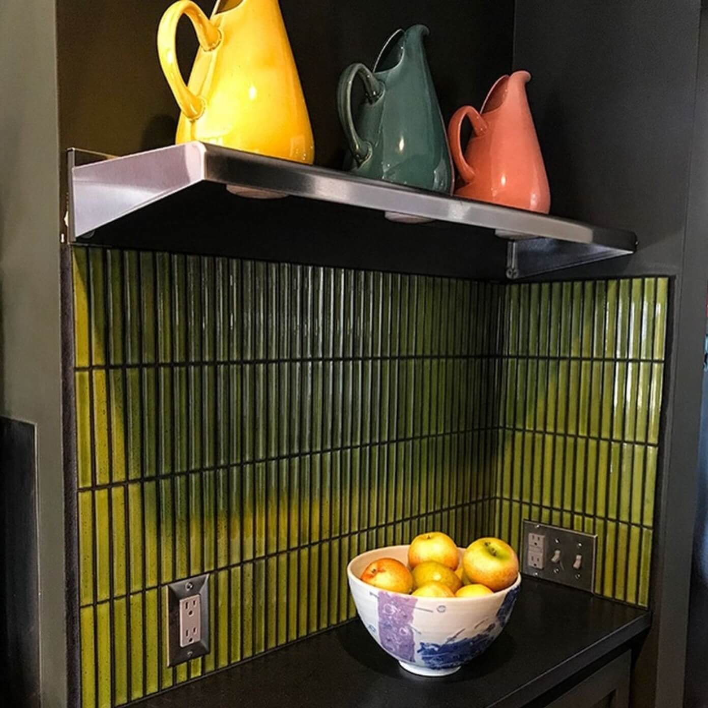 The Sevilla Mosaic Tiles, Original KitKat design in a green Glossy, with a size of 20x145mm installed on a wall on a kitchen with a counter top in front and a fruit bowl containing apple on top. A shelf above holding a yellow, green and red pouring jugs.