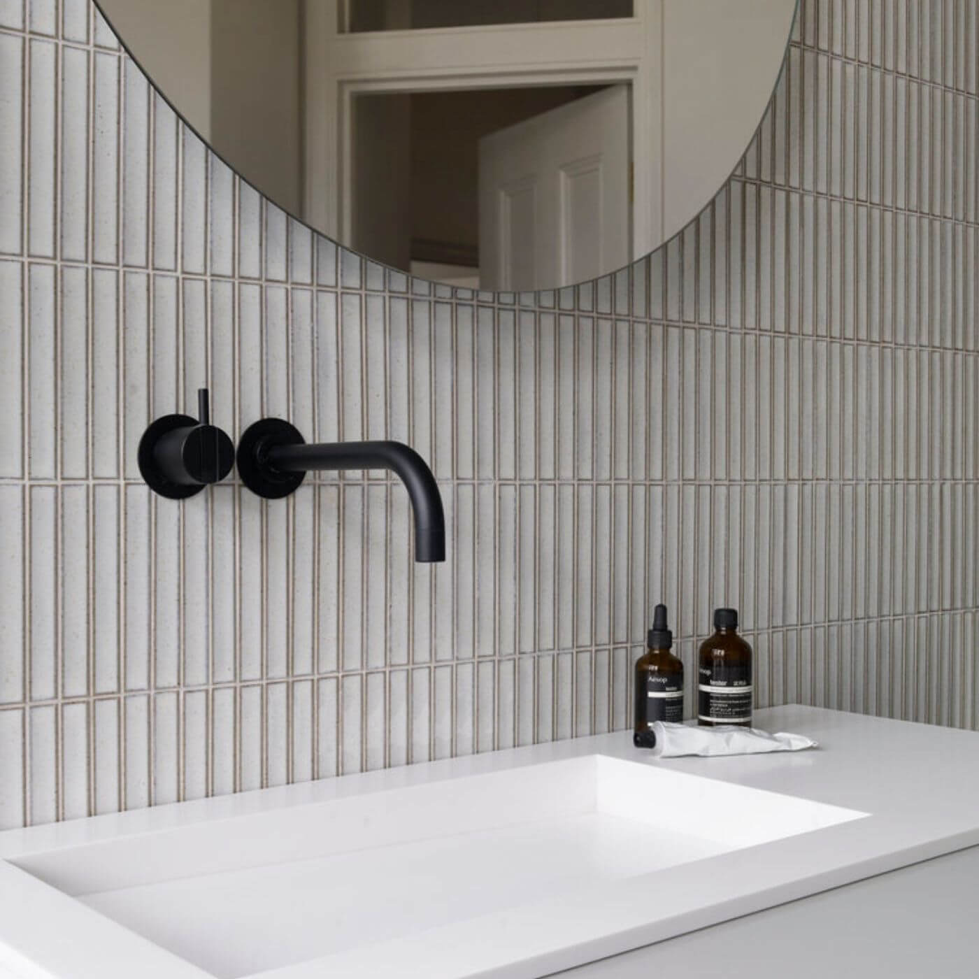 The Sevilla Mosaic Tiles, Original KitKat design in a white blue speckle, with a size of 20x145mm. Installed on a wall in a bathroom behind the vanity unit. A Matt Black tap and mirror fixed to the wall over the top of the tiles.