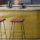 The Sevilla Mosaic Tiles, Original KitKat design in yellow, with a size of 20x145mm. Installed on a kitchen island bench with a stone counter top. Two stools standing in front of the bench. A Blue kit kat style mosaic tile in installed on the wall in the background.